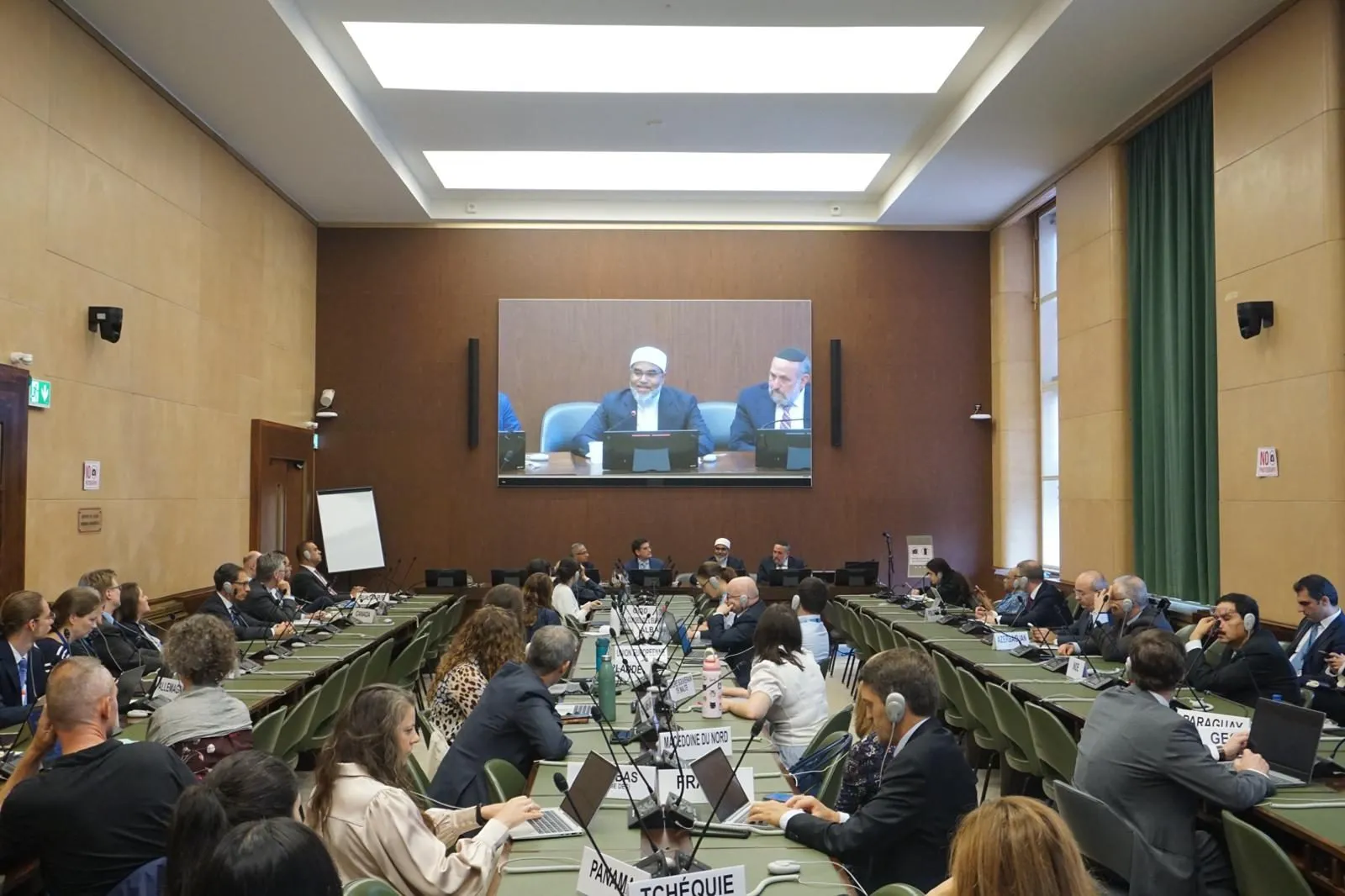 Wide view of the conference room during the side event, with participants at the tables and the panel on screen.