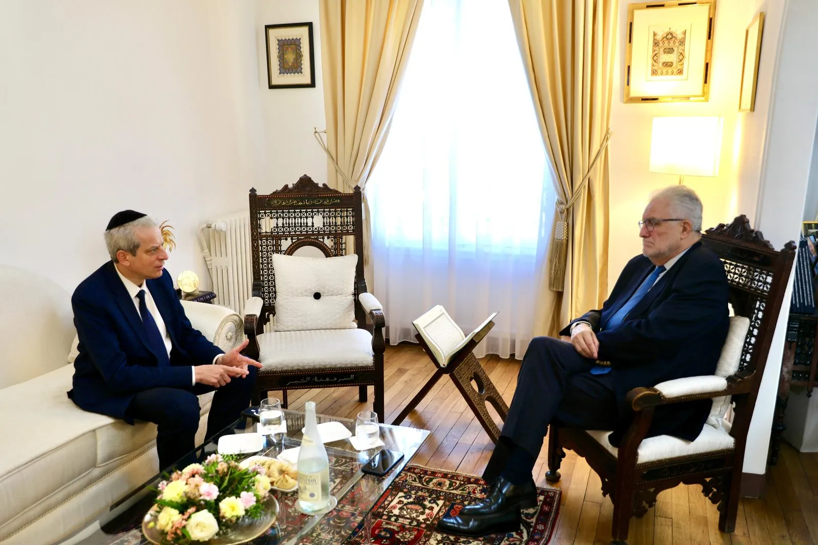 Chief Rabbi Moshe Lewin seated on a sofa and Rector Chems-Eddine Hafiz seated in a carved wooden armchair, in conversation in a reception room.